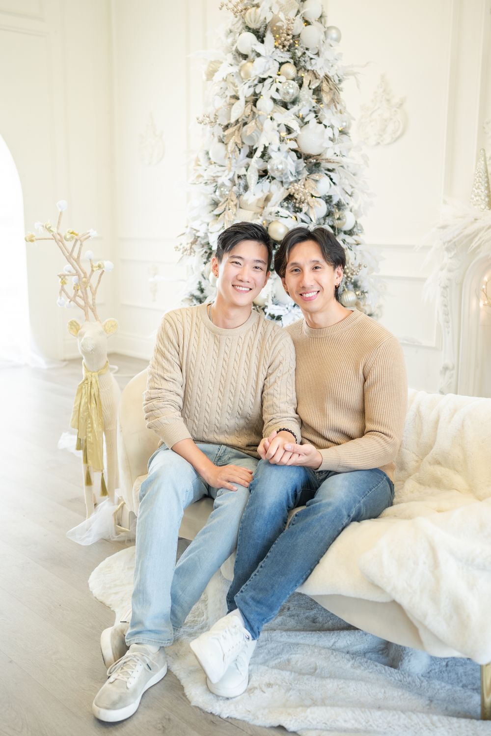 Two people in neutral sweaters sit together on a white sofa with a decorated Christmas tree behind them.