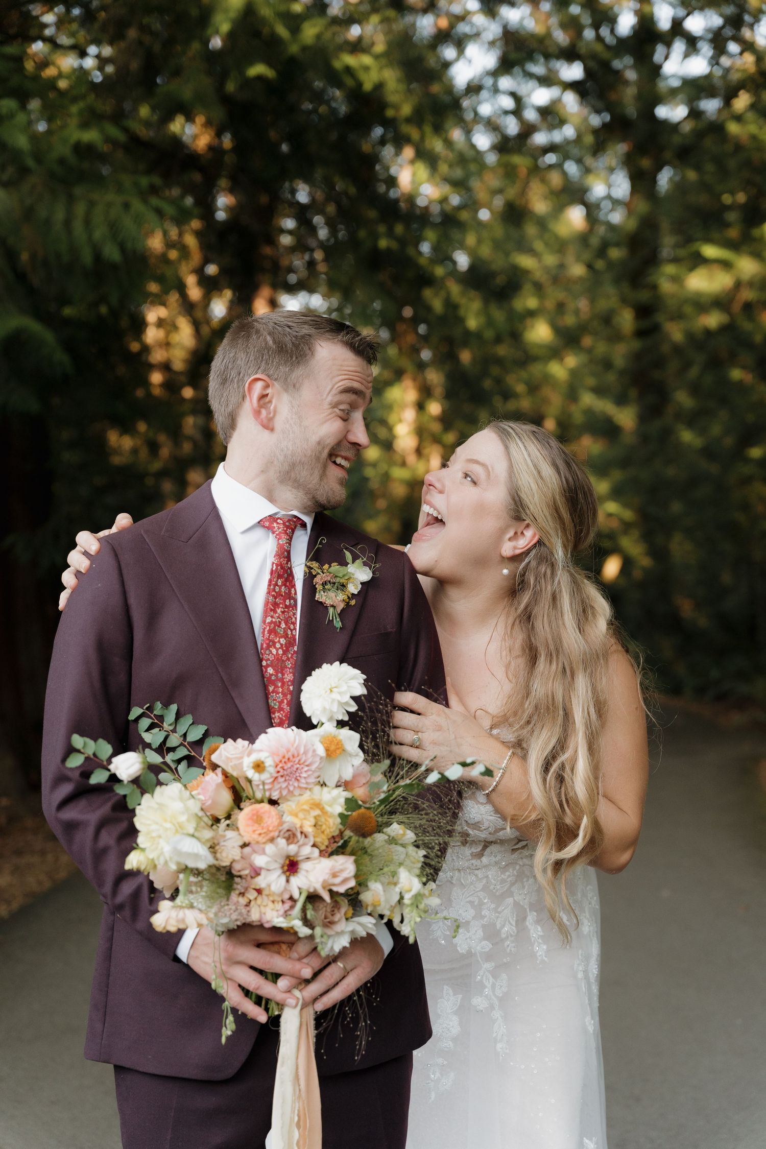 A couple shares a joyful laugh while holding a peach and blush floral bouquet during their outdoor wedding.