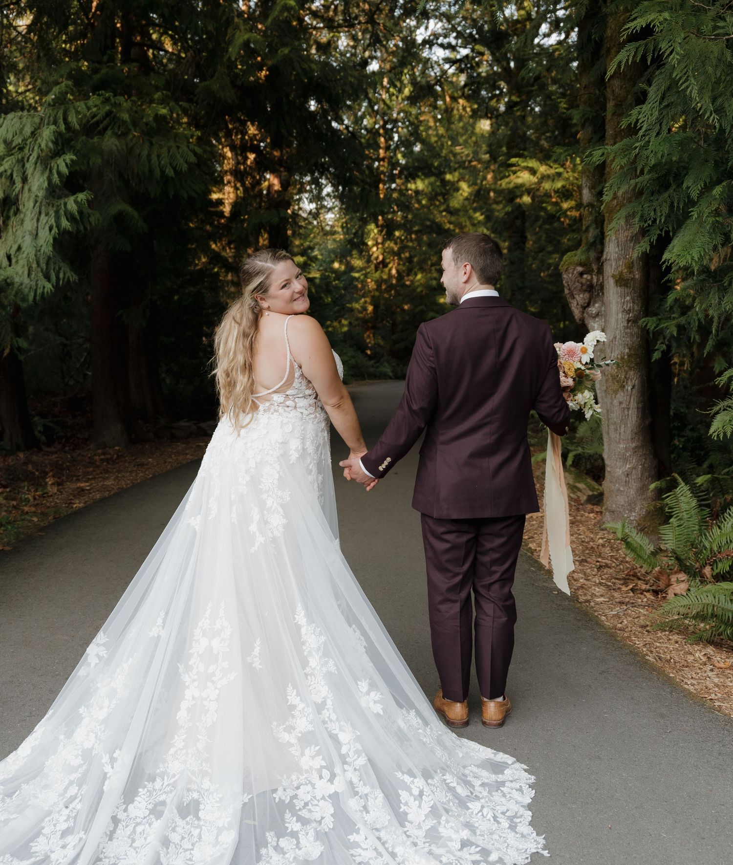 A couple in wedding attire walk hand in hand down a tree-lined path, with the bride's lace train trailing behind.