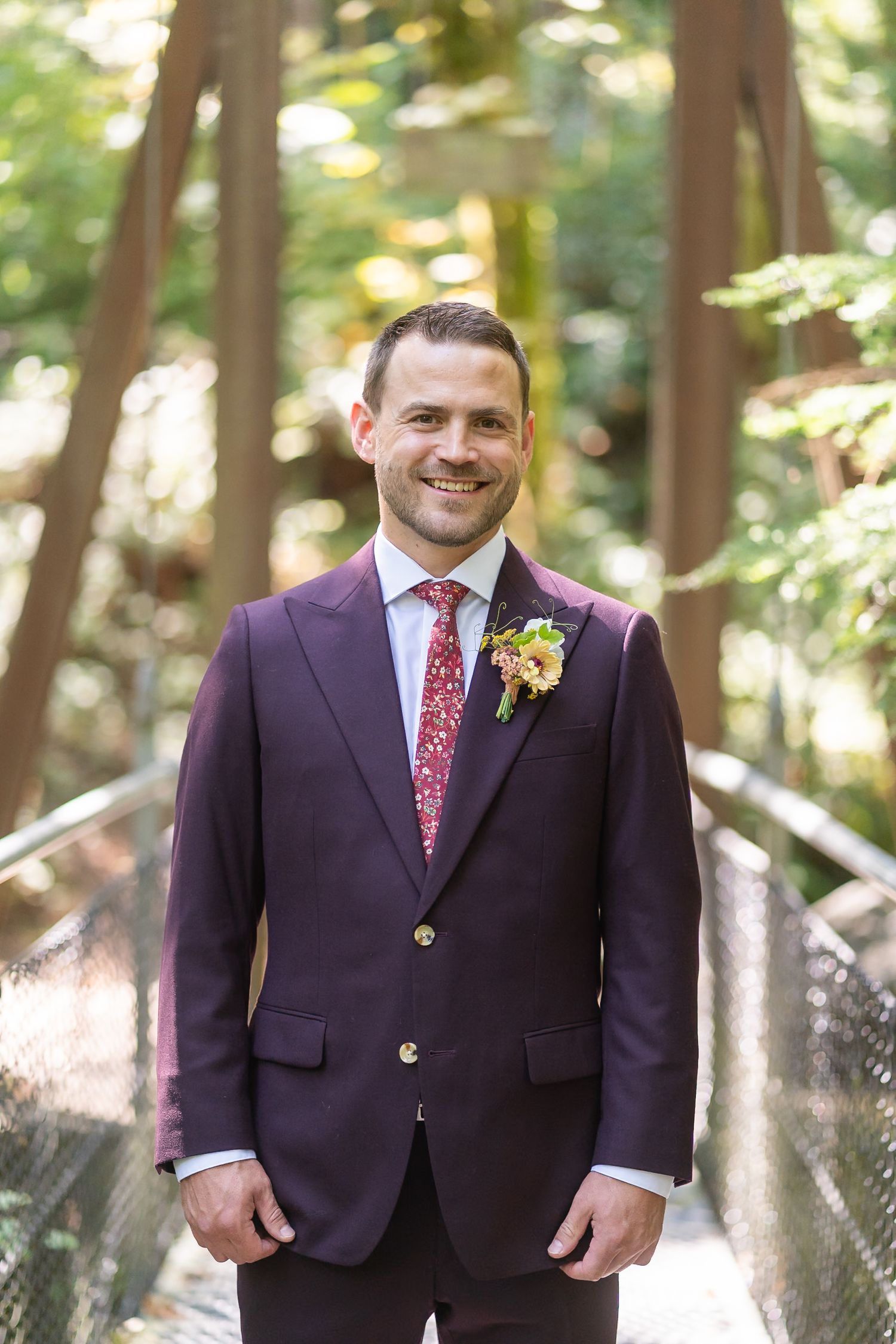 A groom in a purple suit with red patterned tie and floral boutonniere stands in a forest setting at a wedding.