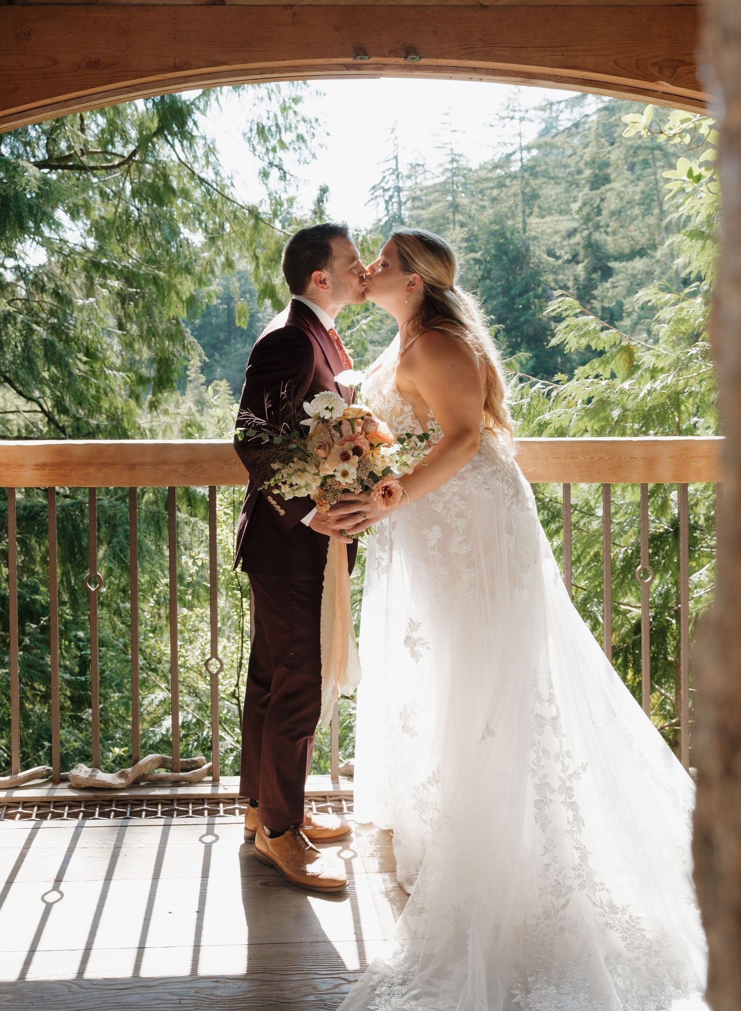 A couple shares their first kiss as newlyweds on a wooden deck surrounded by evergreen forest scenery.