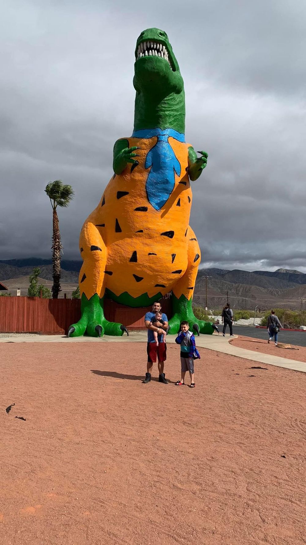 T-Rex dinosaur display in California with orange body and green details like Flinstones towers over visitors at an outdoor attraction.