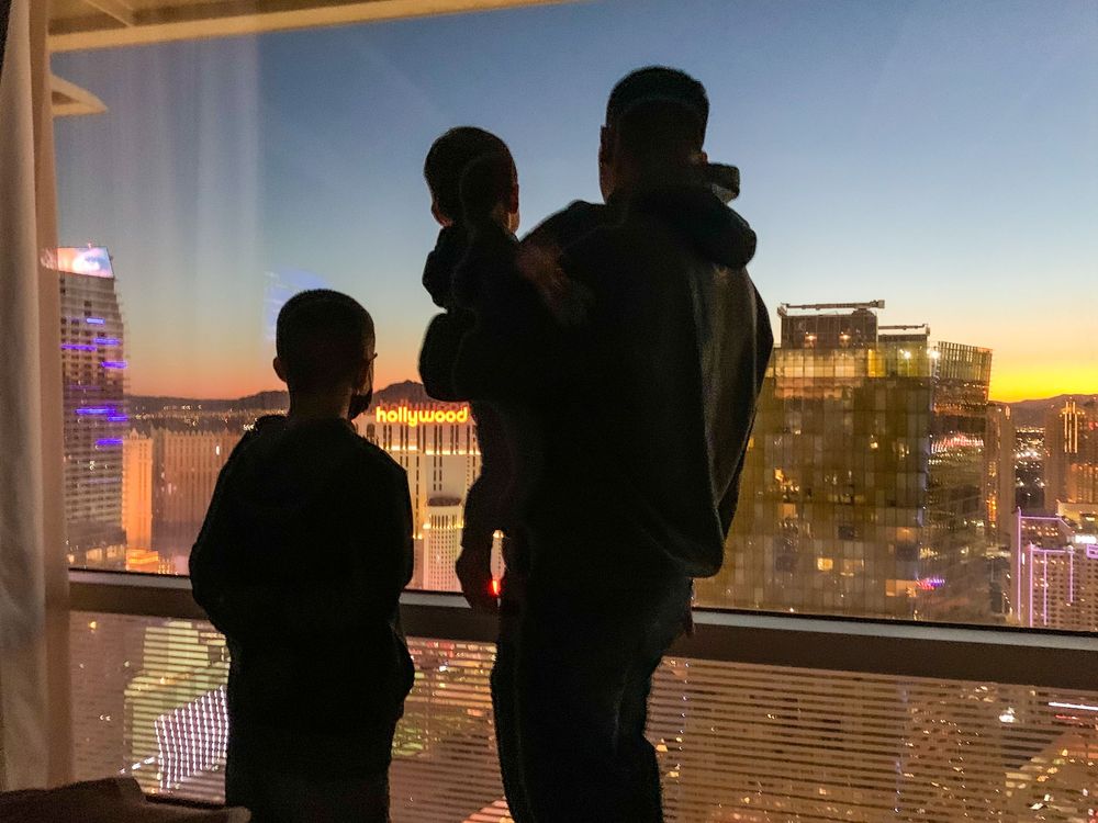 Silhouettes stand at a window overlooking a city skyline during sunset with illuminated buildings in the background.