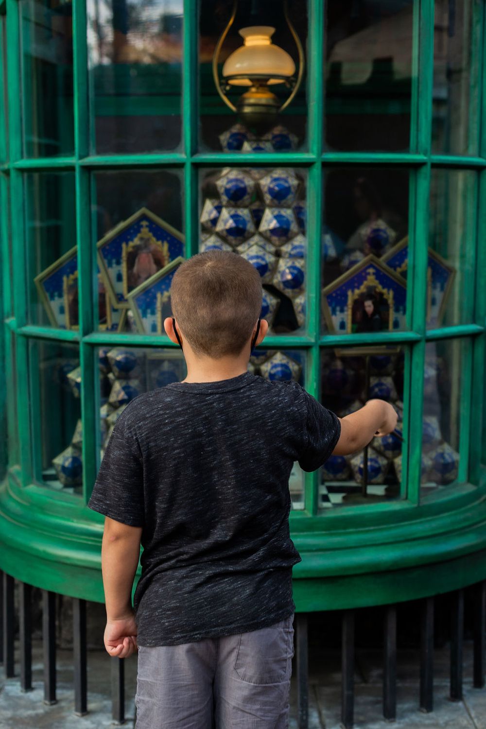 A child looks through a green-framed window display at blue collectible merchandise in The Wizarding World theme park.