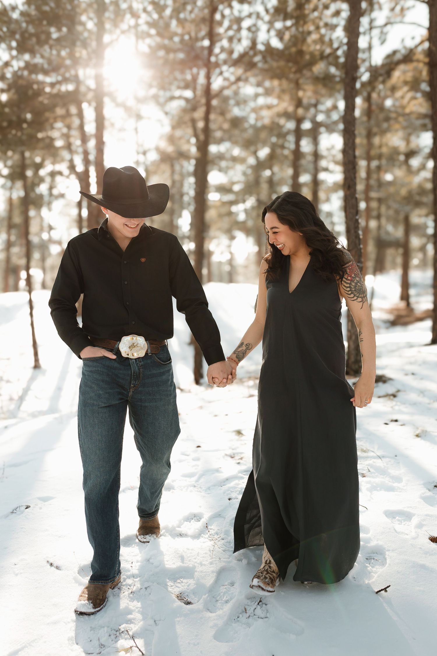 A couple walks hand in hand through a snowy forest path with sunlight streaming through pine trees in winter.