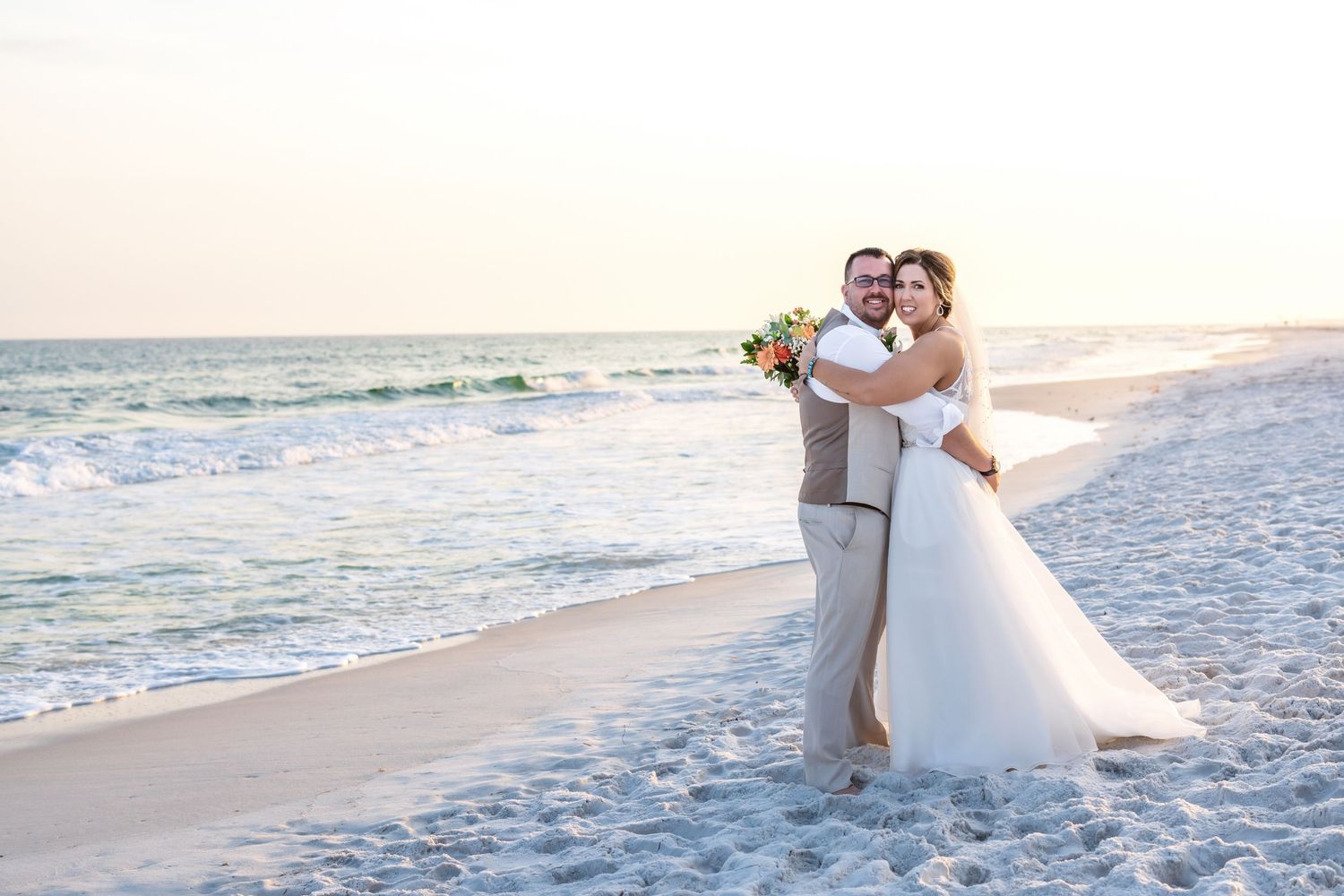 A newlywed couple embraces on a white sandy beach at sunset with gentle waves in the background.