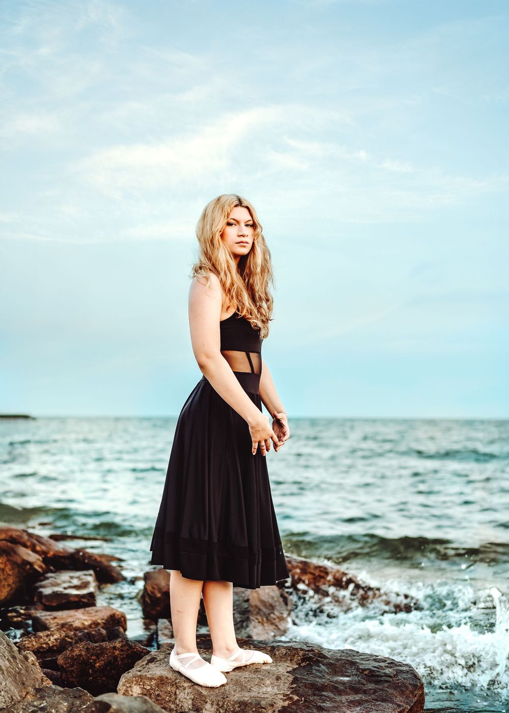 A person in a black ensemble stands on coastal rocks by the ocean waves wearing white shoes.