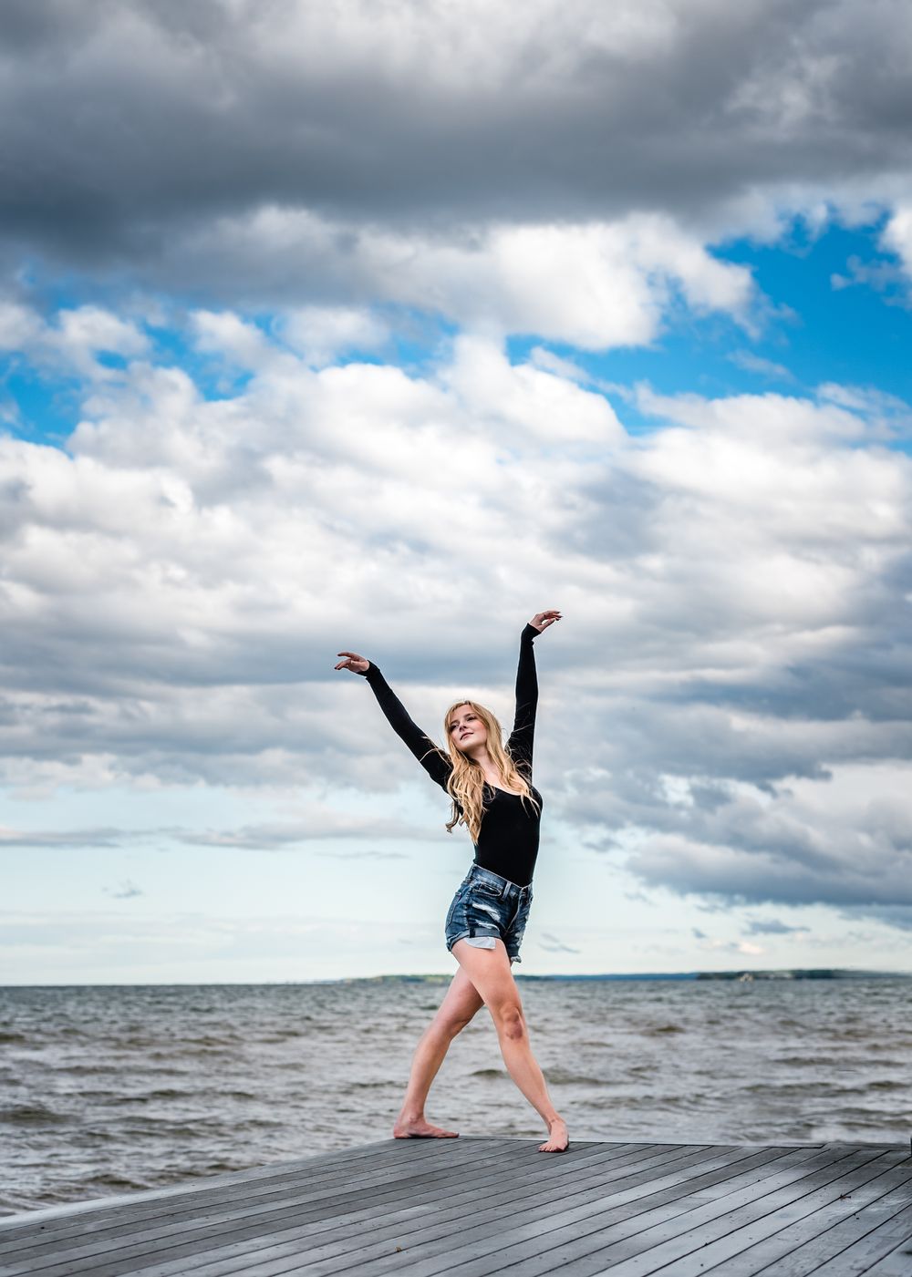 A joyful figure dances on a wooden pier overlooking the ocean under dramatic cloudy skies.