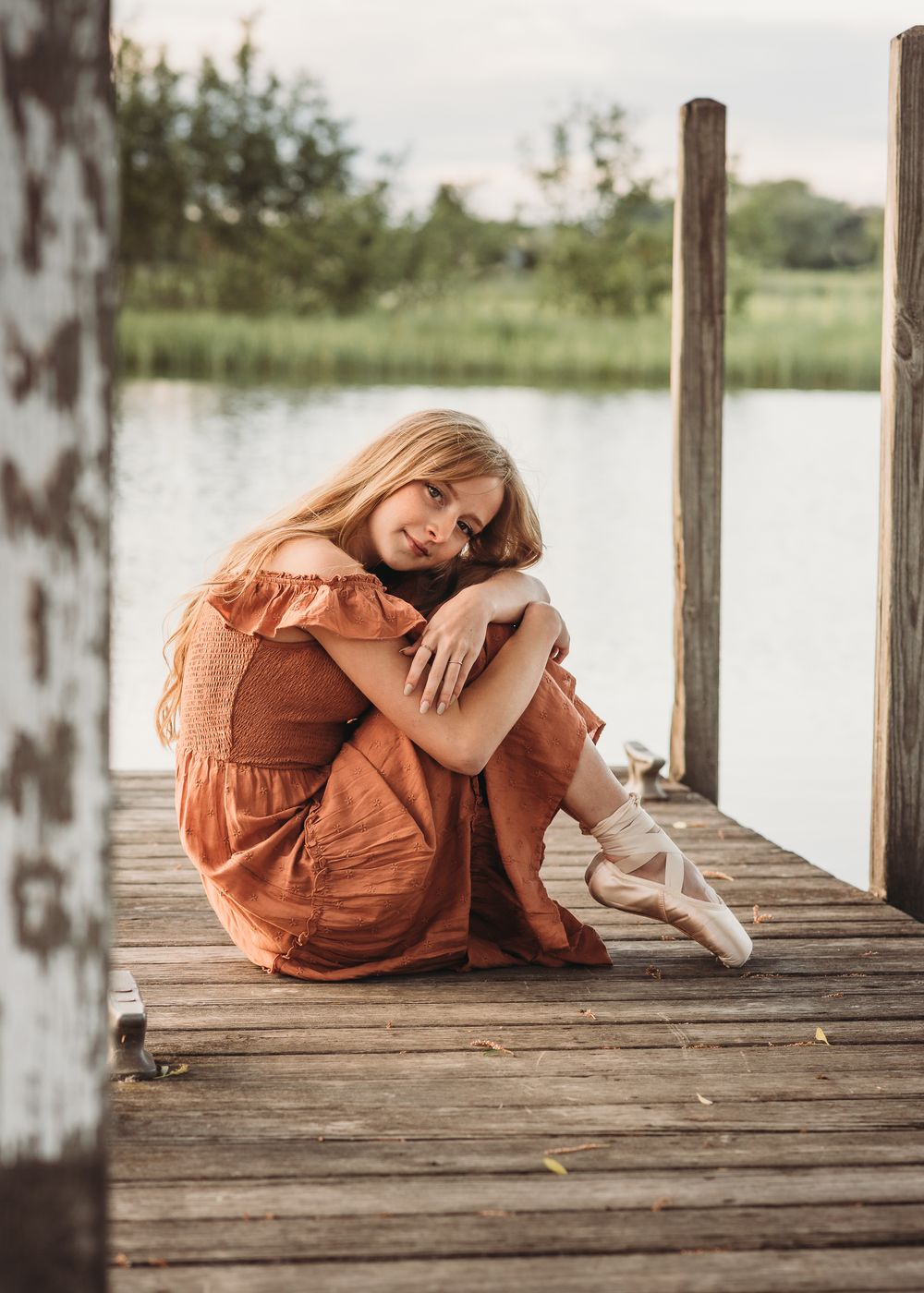 A rustic wooden dock setting featuring ballet shoes and a flowing rust-colored dress against a serene lake backdrop.