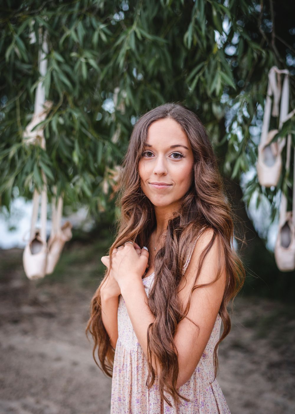 A woman with very long wavy brown hair poses in a light dress under a willow tree with hanging decorations.