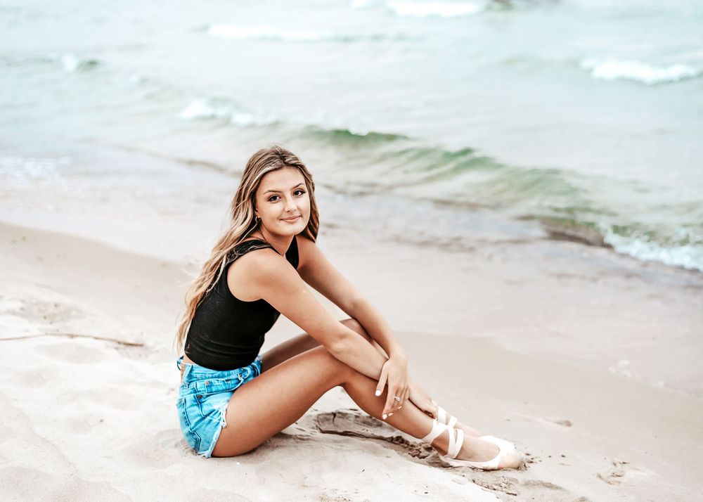 Person in black tank top and blue patterned shorts sitting on a sandy beach with waves in the background.