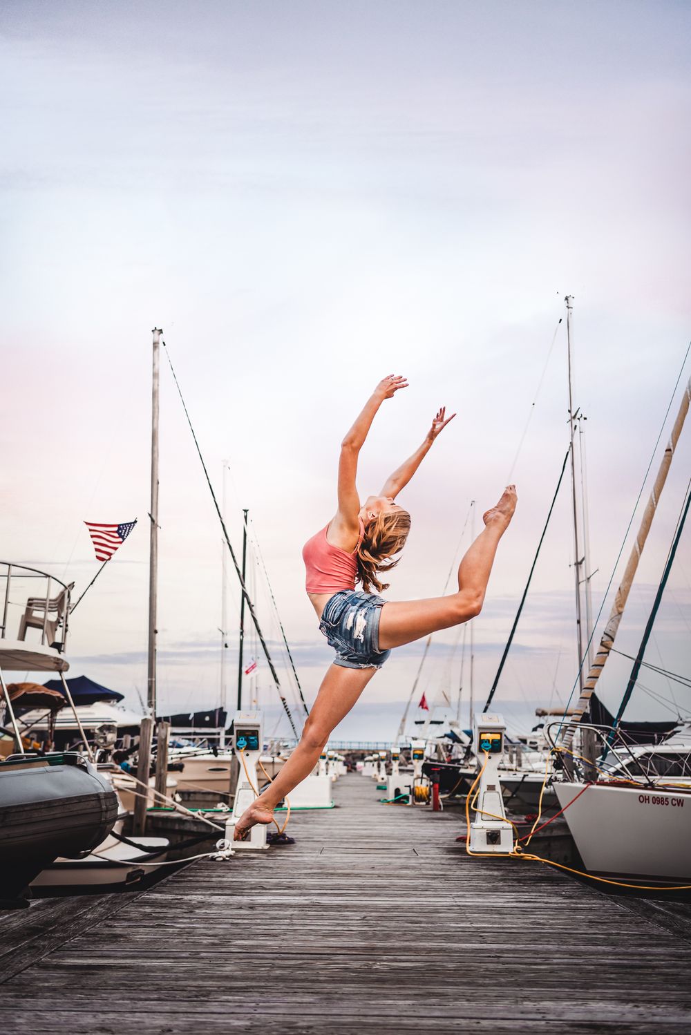 A dancer performs a graceful leap on a wooden dock surrounded by sailboats and yachts in a marina at sunset.