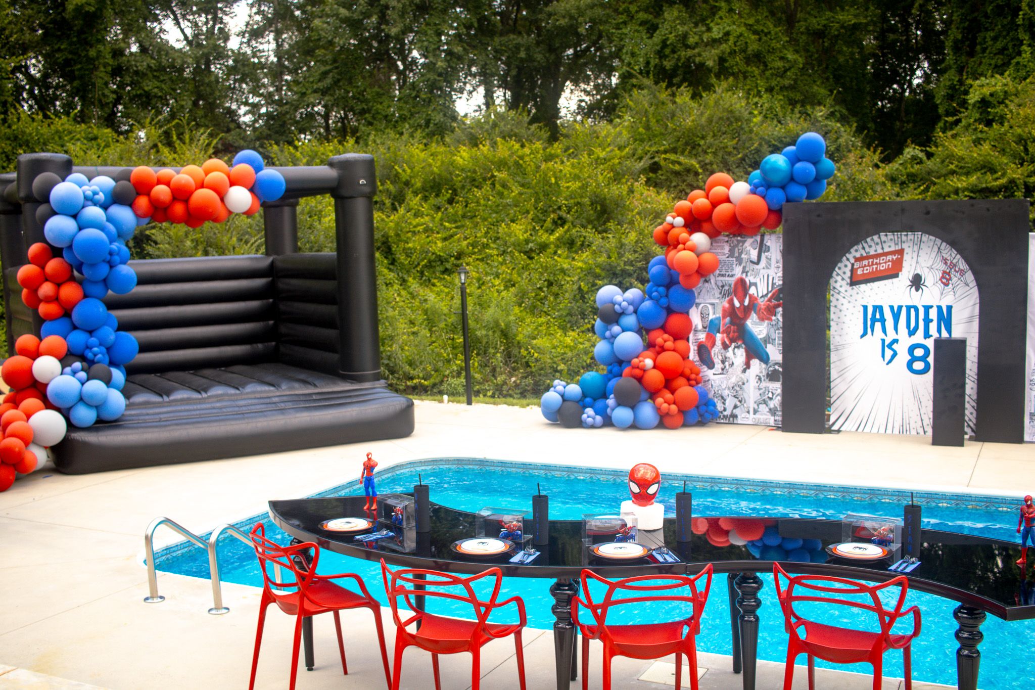 Spider-Man themed poolside party setup with red and blue balloon arch decorations and red chairs.