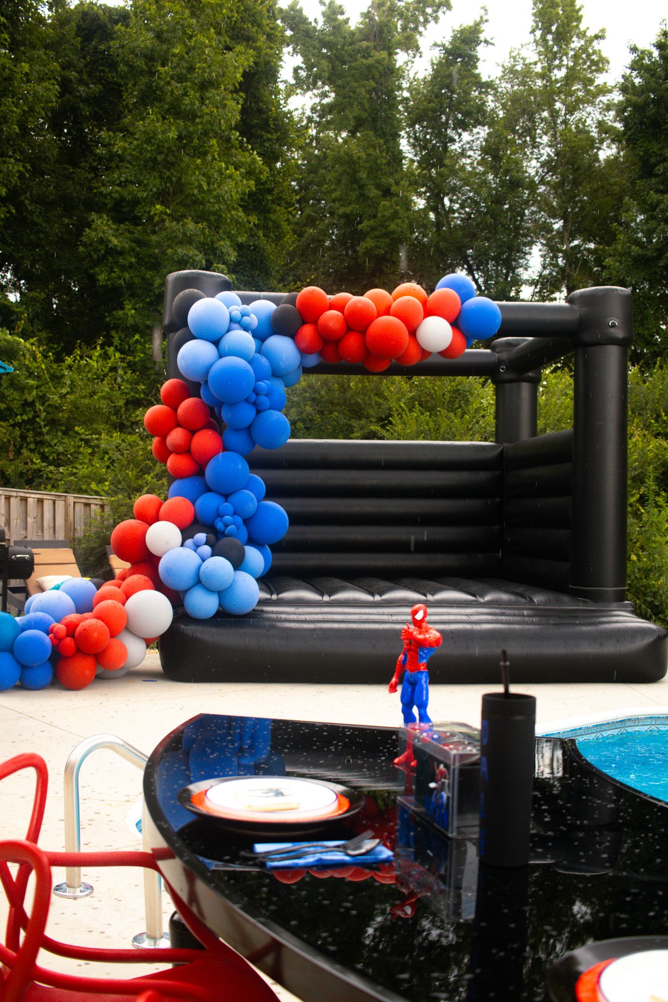 Spider-Man themed bouncy castle decorated with red and blue balloon garland beside a pool.