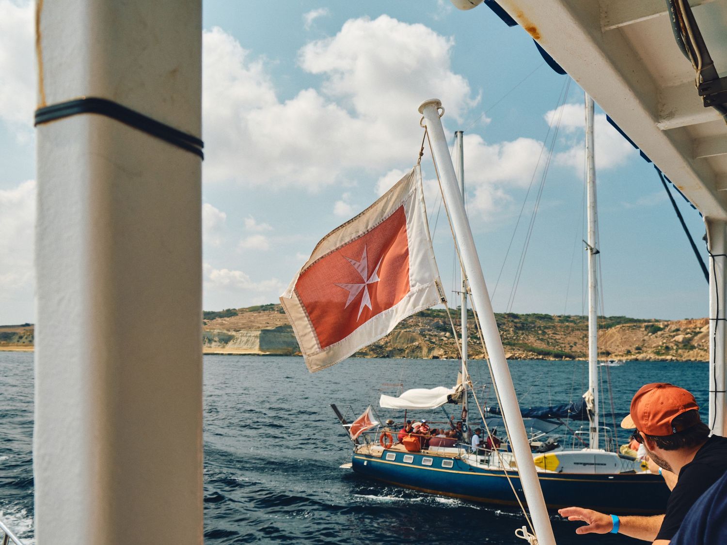 Red flag waves on a sailboat mast against blue waters and coastal cliffs on a sunny day at sea.
