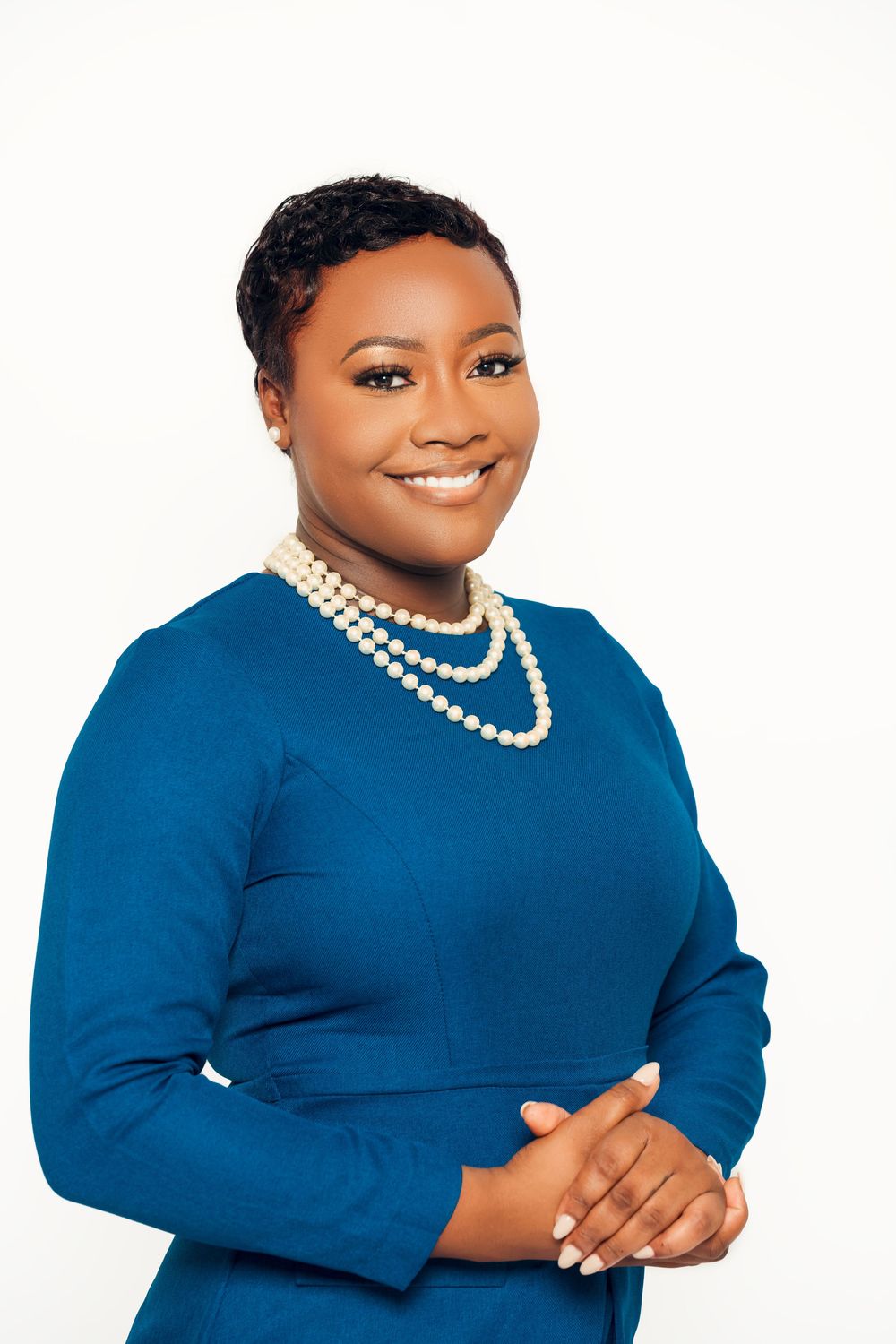 Professional portrait of someone in a royal blue dress with layered pearl necklaces against a white background.
