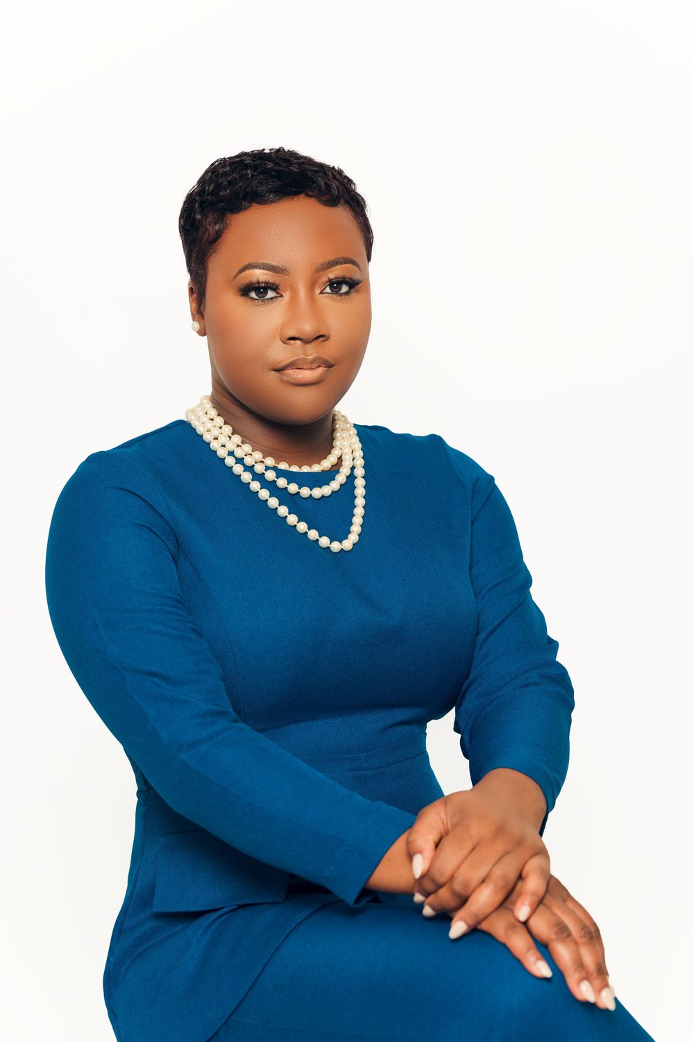 Professional portrait of a person wearing a blue dress with long sleeves and a layered pearl necklace against white background.
