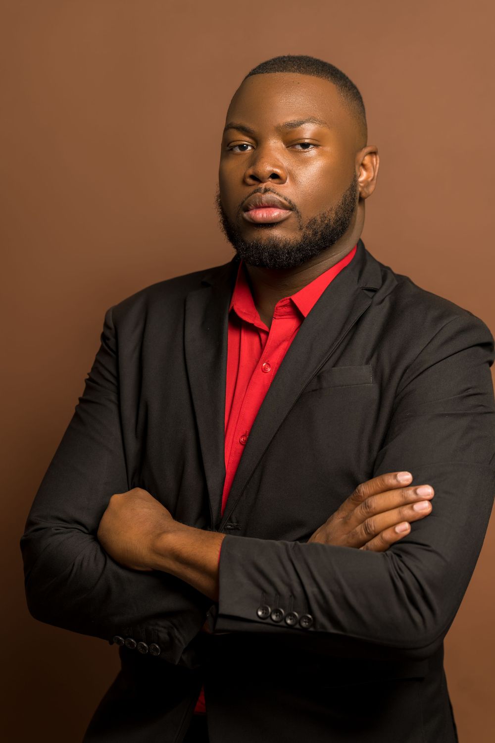 Professional studio portrait of someone in a black suit jacket and red dress shirt against a brown background.