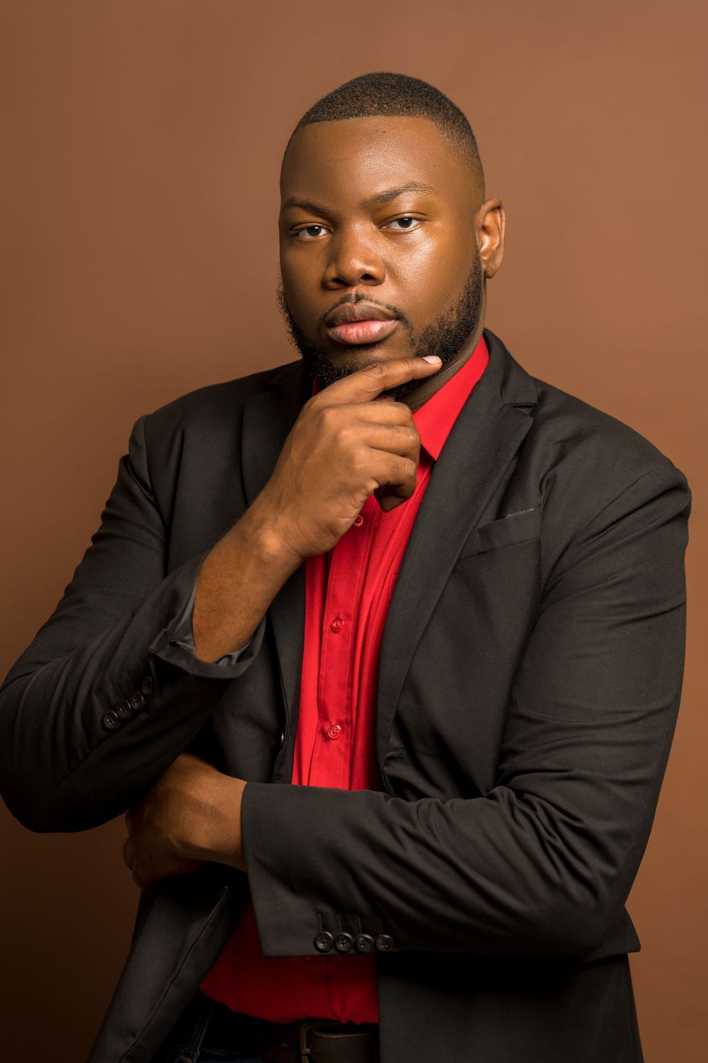 Professional portrait of someone in a black blazer and red button-up shirt striking a thoughtful pose against brown backdrop.