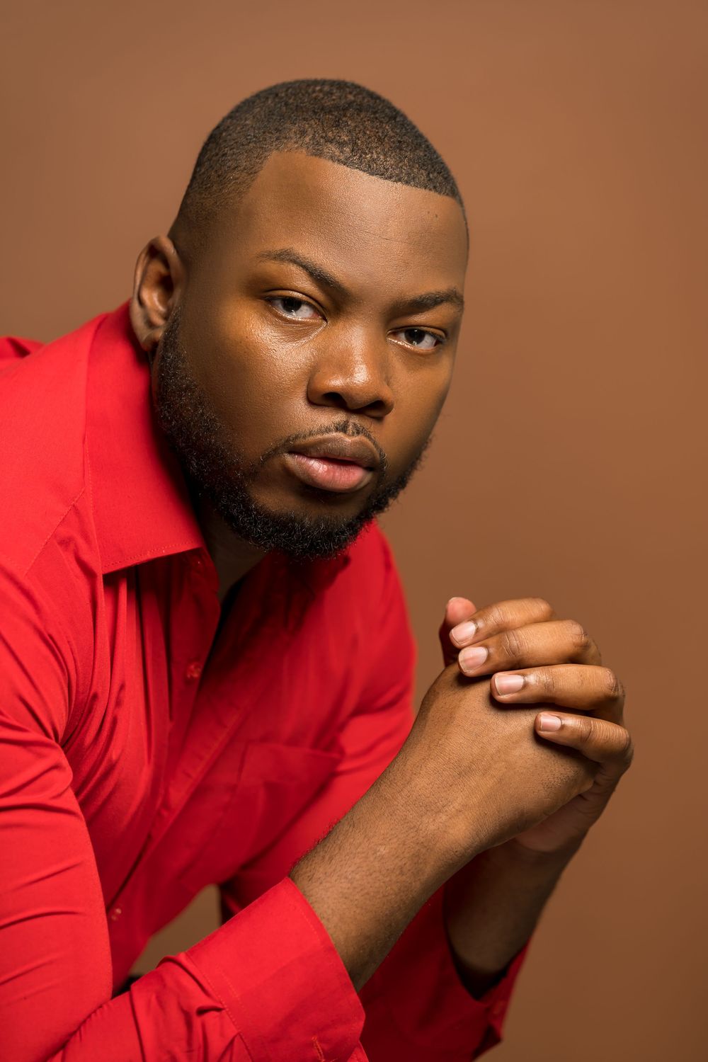 Portrait against brown background features person in red long-sleeved shirt with clasped hands in contemplative pose.