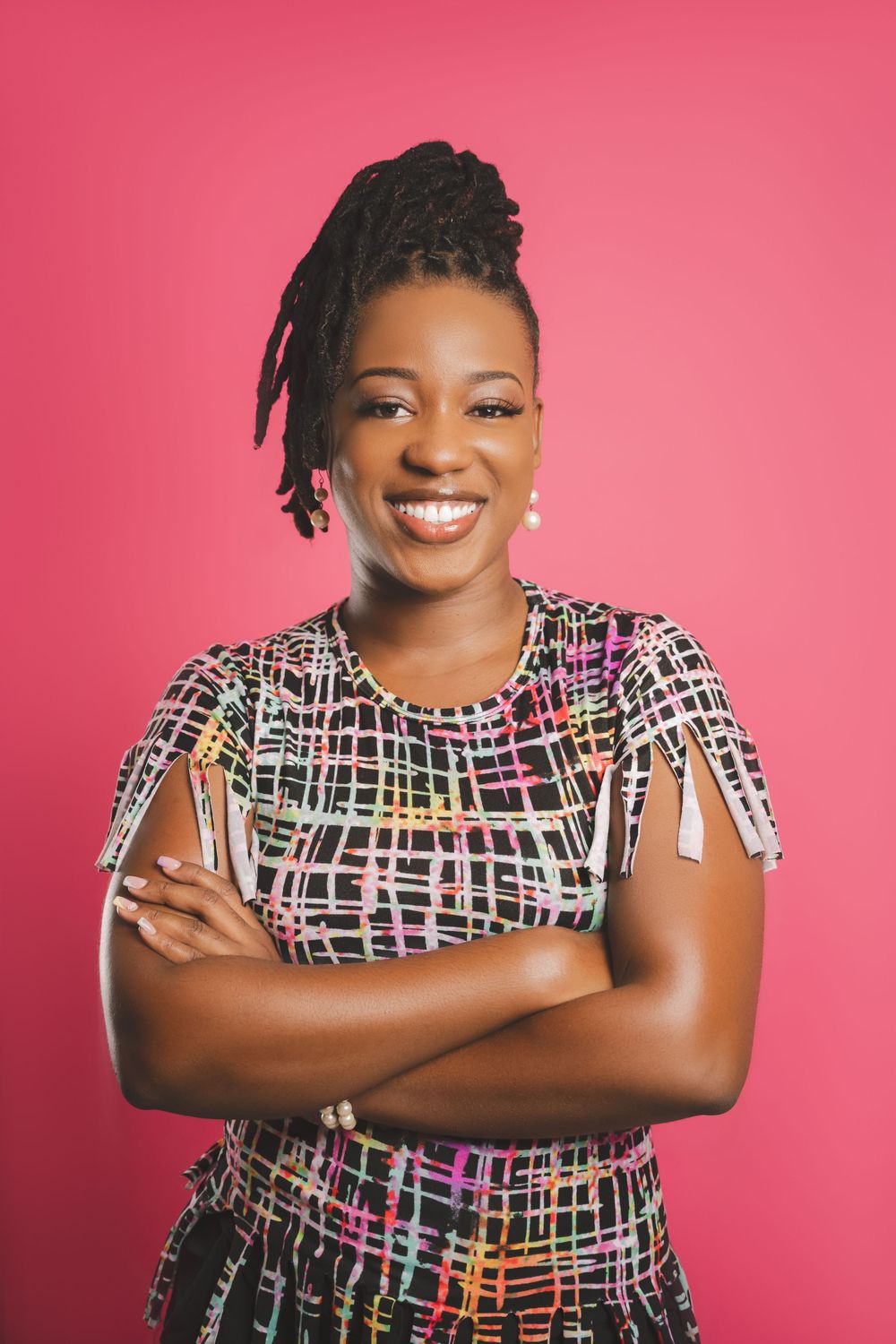 A person wearing a colorful patterned dress stands confidently with crossed arms against a pink background.