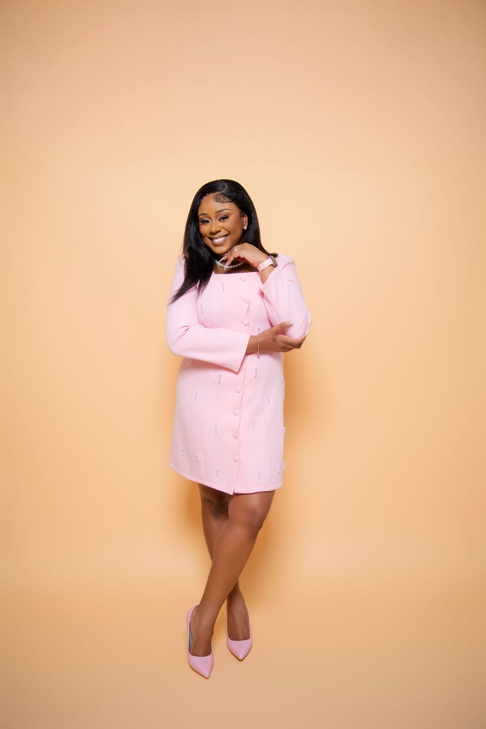 A woman wearing a pink button-down shirt dress and matching heels poses confidently against a beige backdrop.