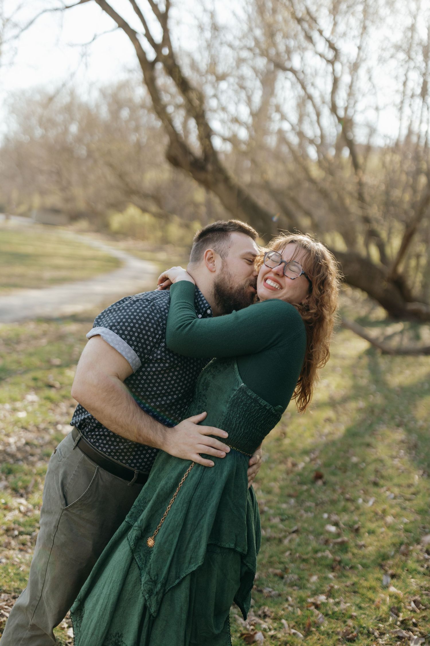 Romantic couple in green outfits share tender embrace while laughing together outdoors among bare trees in autumn.