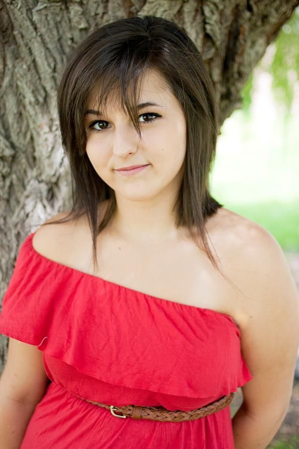 A young woman in a bright red off-shoulder dress with a brown belt poses outdoors against a tree trunk.