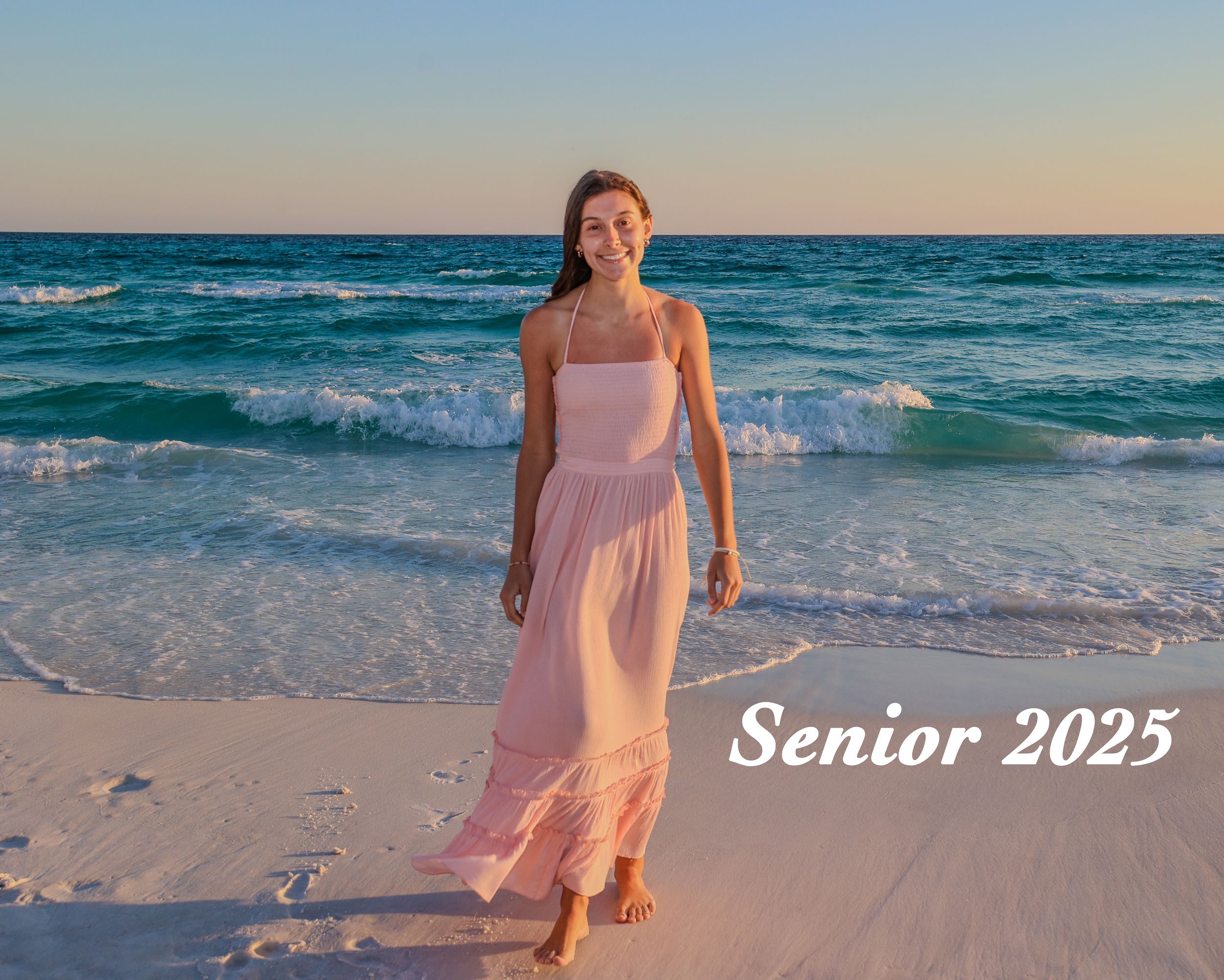 A graduating senior in a flowing pink dress walks along a beach at sunset with turquoise waves crashing behind her.