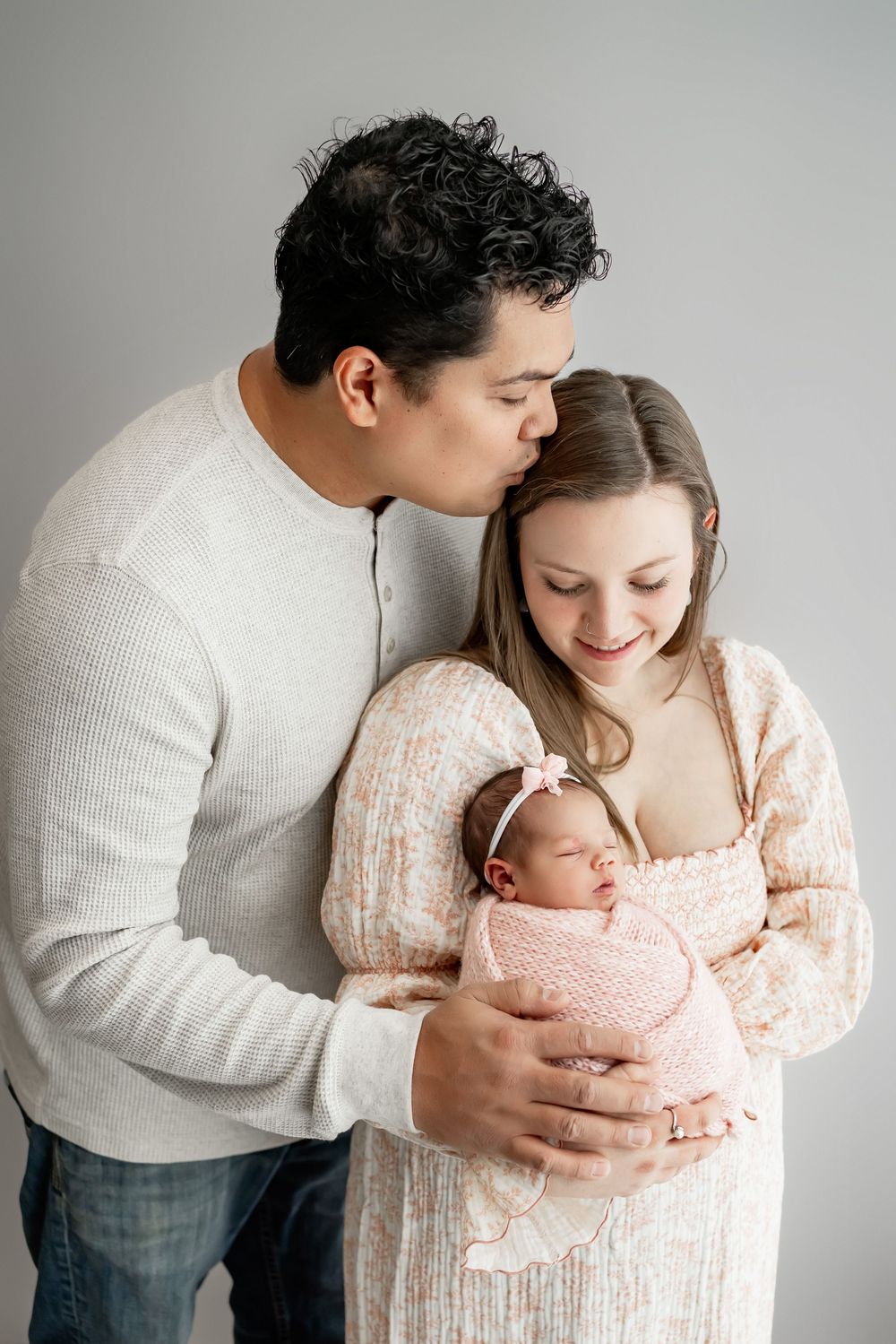 Family in white attire sharing tender moments with newborn baby against neutral backdrop.