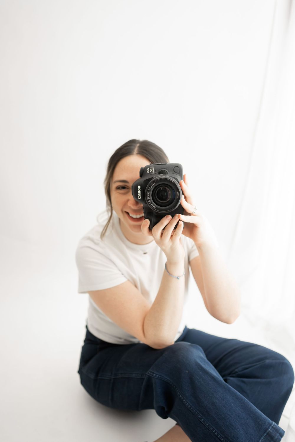 Photographer in white t-shirt holds professional camera in minimalist white studio setting.