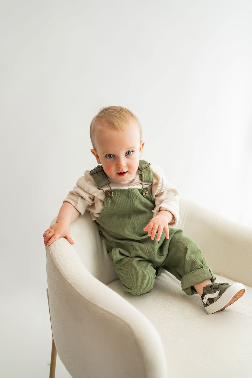 Baby in olive green overalls sits in white modern chair.