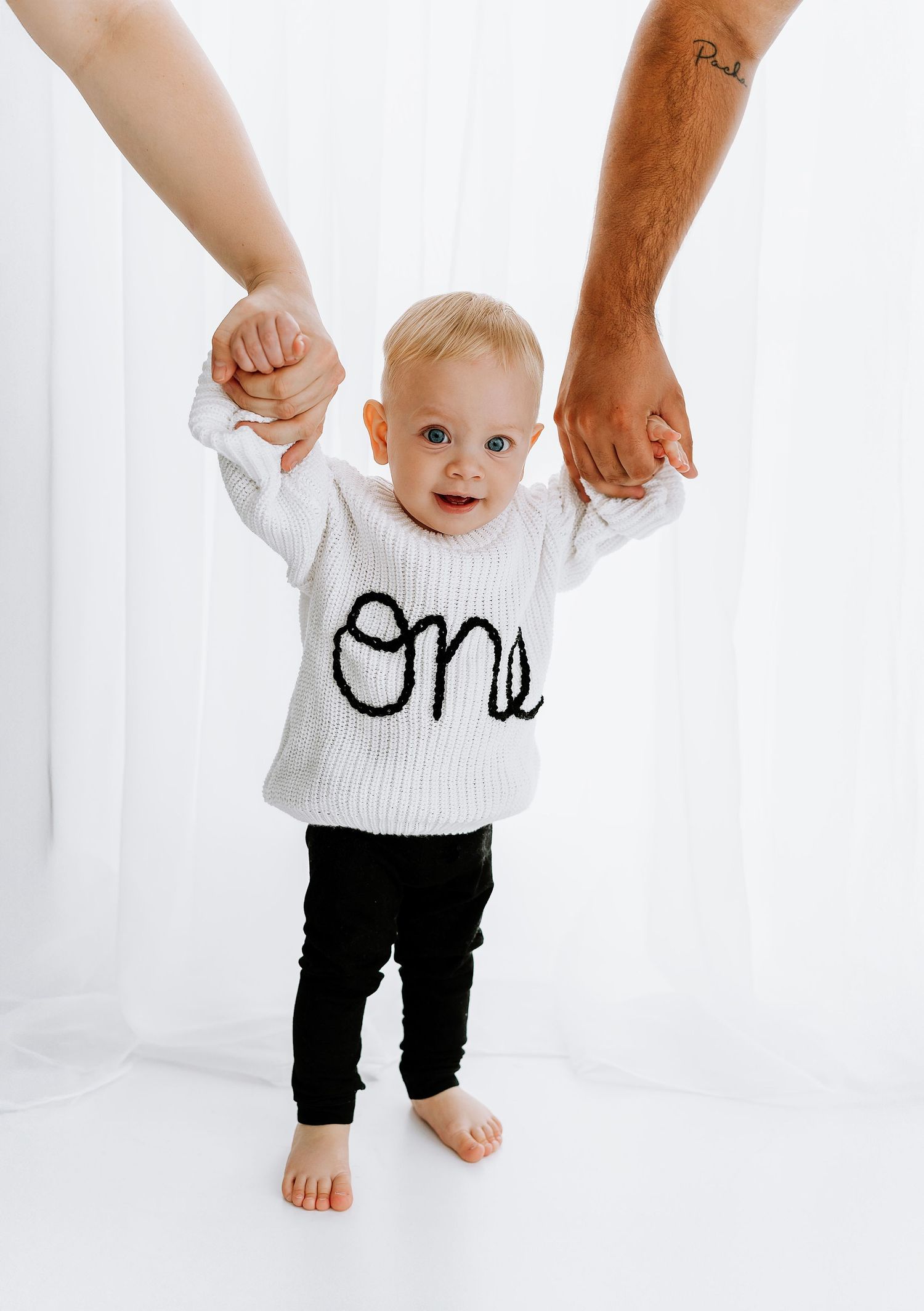 A baby in a white sweater with 'One' written on it holds hands with adults while standing barefoot against a white backdrop.