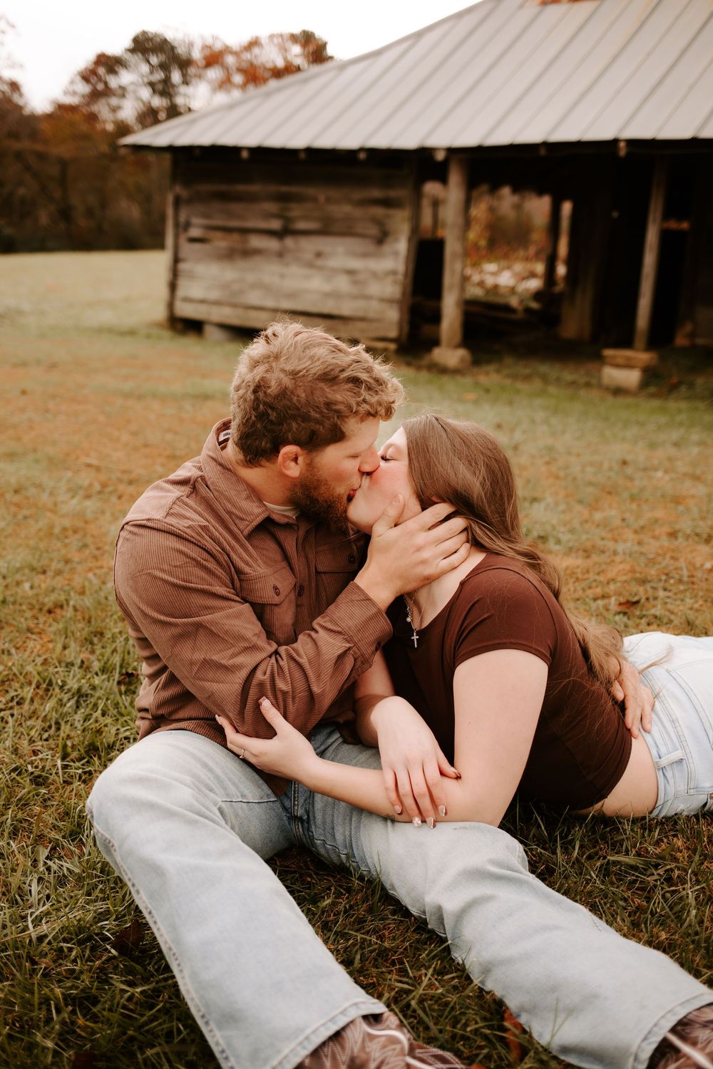A couple shares a romantic kiss while sitting on grass in front of a rustic wooden barn during fall.