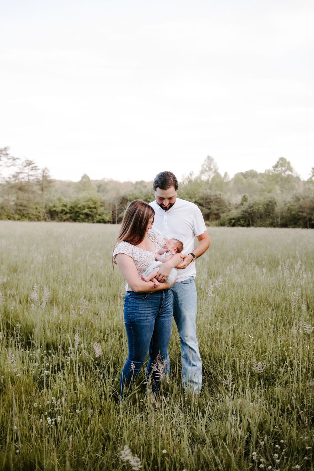 New parents lovingly gazing at their newborn baby while standing in a lush green meadow at dusk.