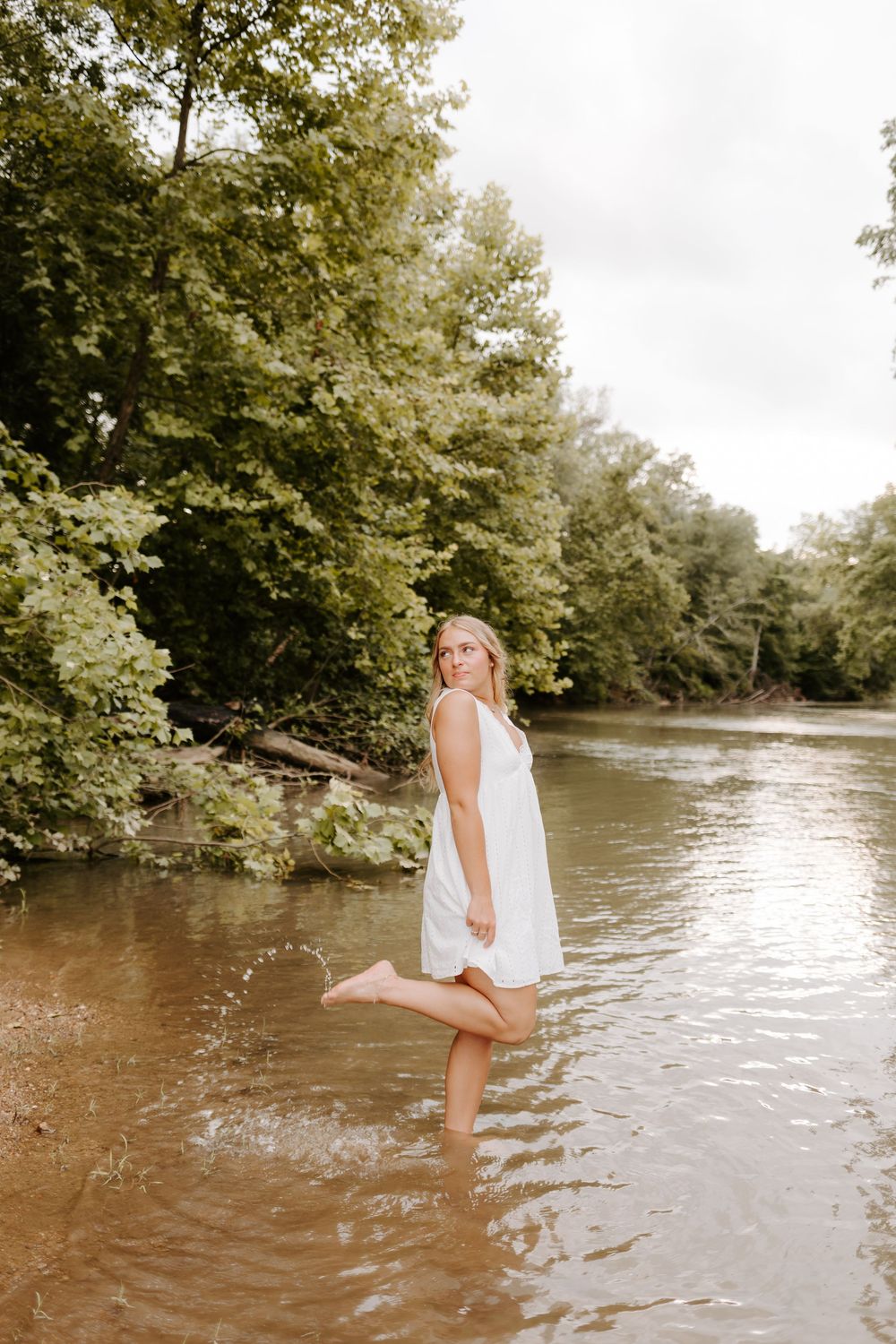 Young blonde woman in a white sundress standing in a shallow river surrounded by lush green trees at Ketner's Mill, Whitwell Tennessee