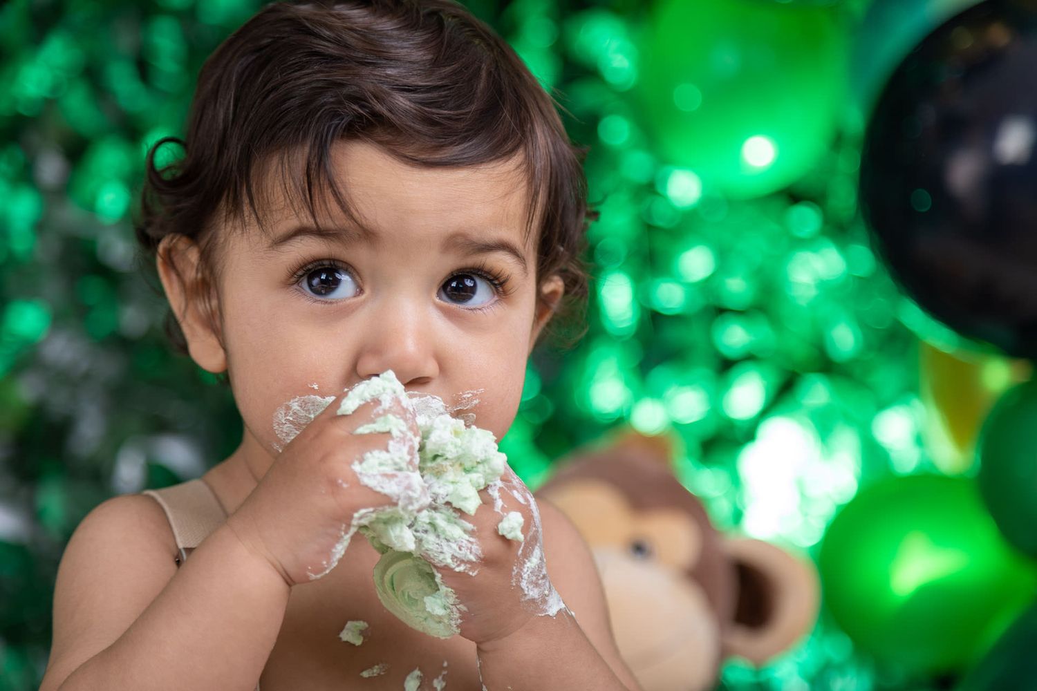 Baby smashing cake into face with green balloons and lights in background at birthday party celebration.