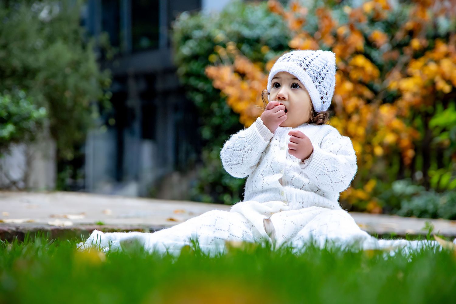 Baby in white knit clothing and hat sits on blanket outdoors with autumn foliage in background.