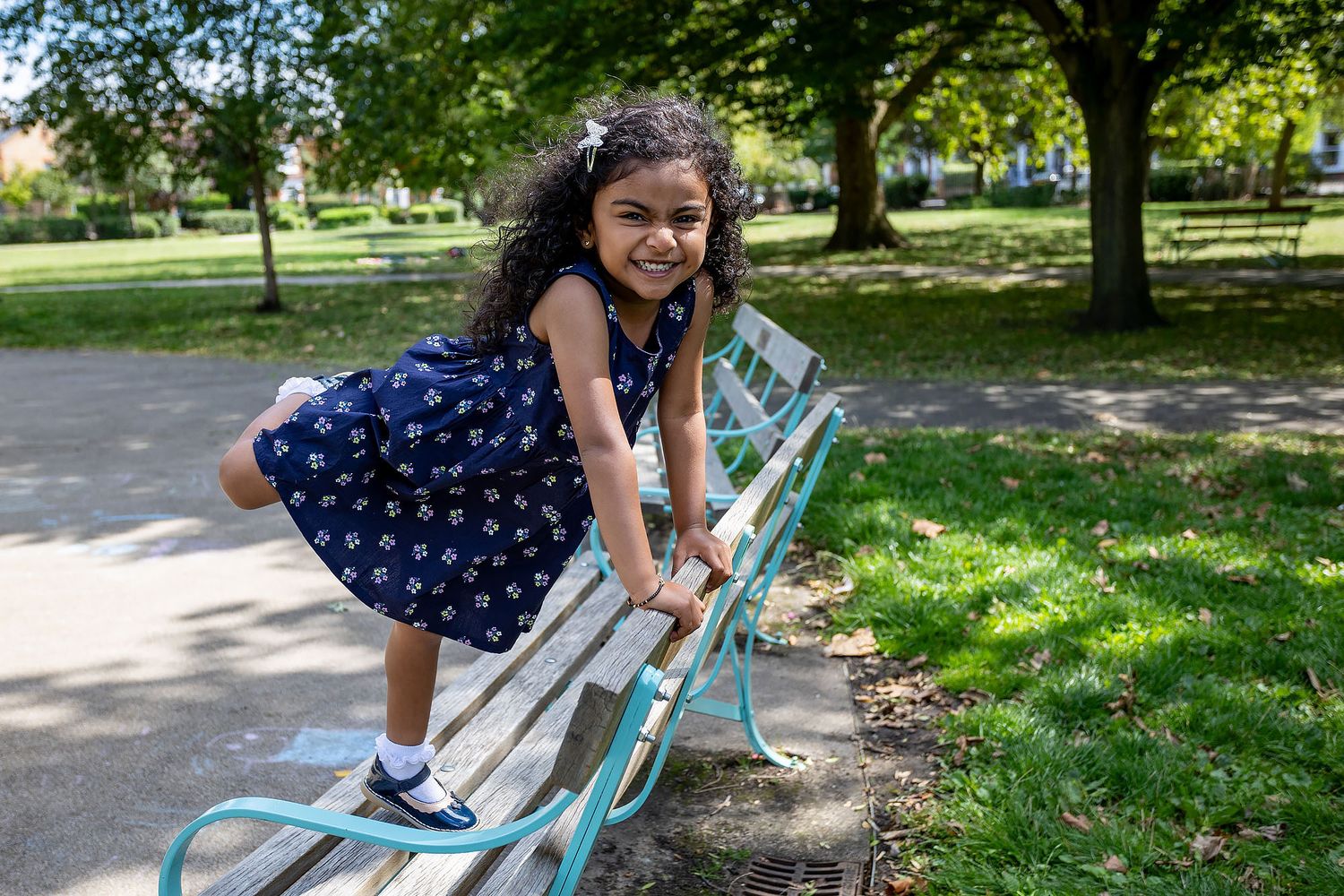 Child playing outdoors leans forward playfully on a park bench while enjoying a sunny summer day in a green space.