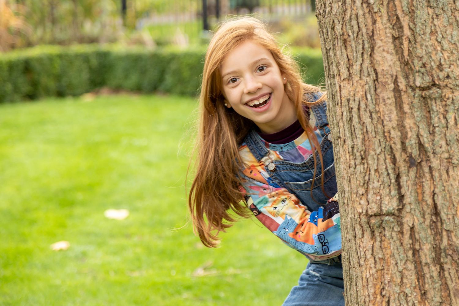A cheerful child with red hair wearing a denim overall peeks out from behind a tree trunk in a sunny garden.