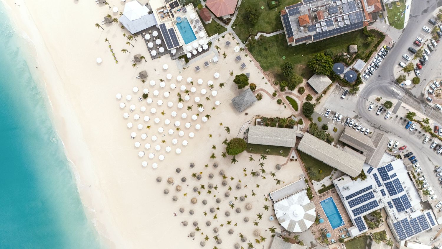 Aerial view of a luxury beachfront resort with turquoise waters, white sand, and beach umbrellas along the coastline.