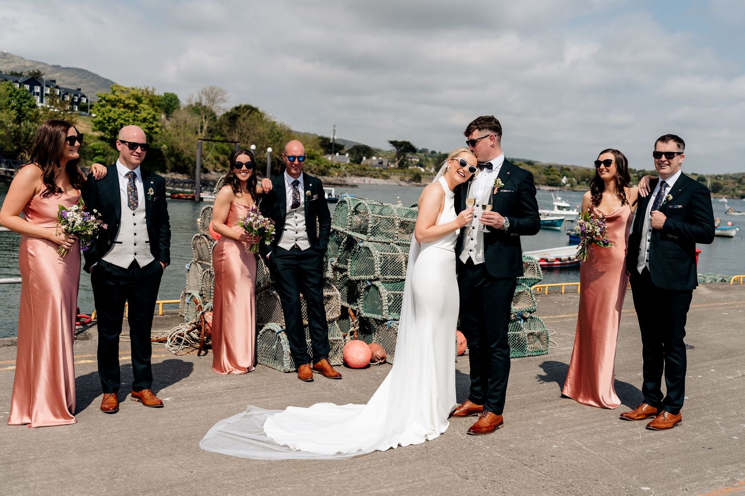 Wedding party in formal attire poses on a sunny harbor dock with boats and hills in the background.