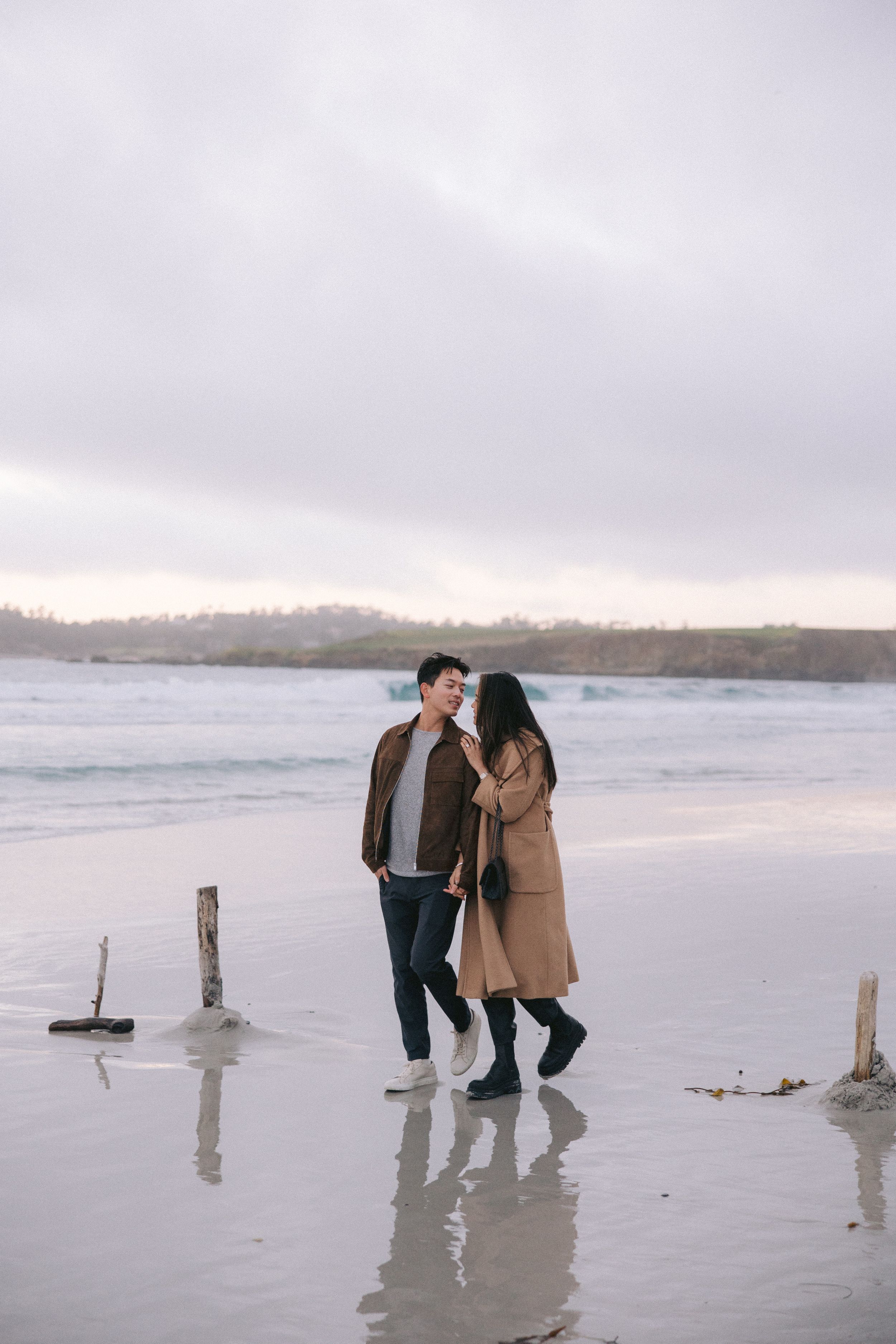 A couple walking together on a misty beach at dusk with cloudy skies and wooden posts in the sand.