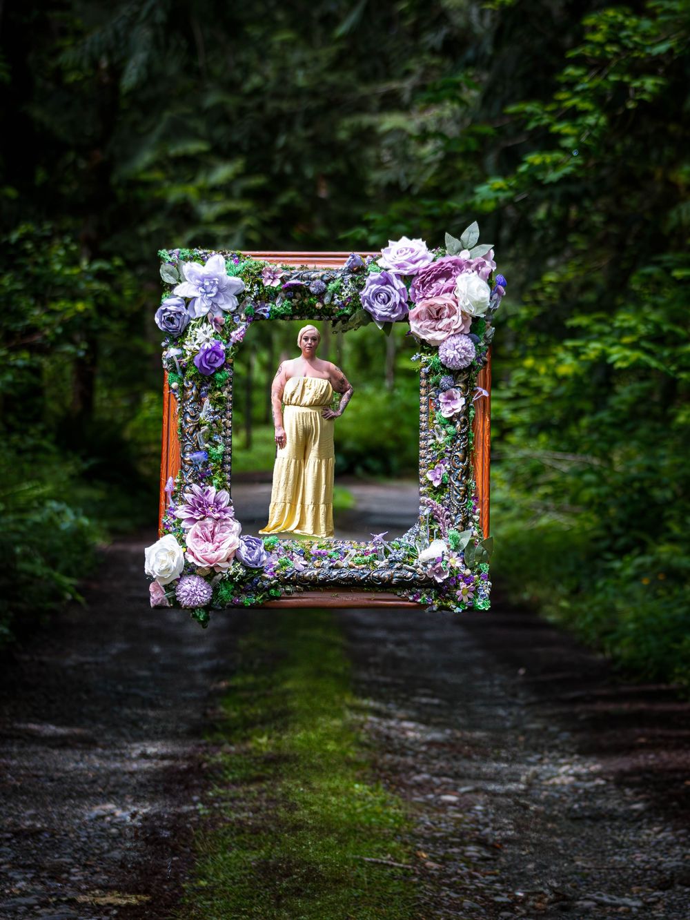 A decorative wooden frame adorned with purple and pink flowers stands on a forest path creating a magical garden scene of a woman in yellow.