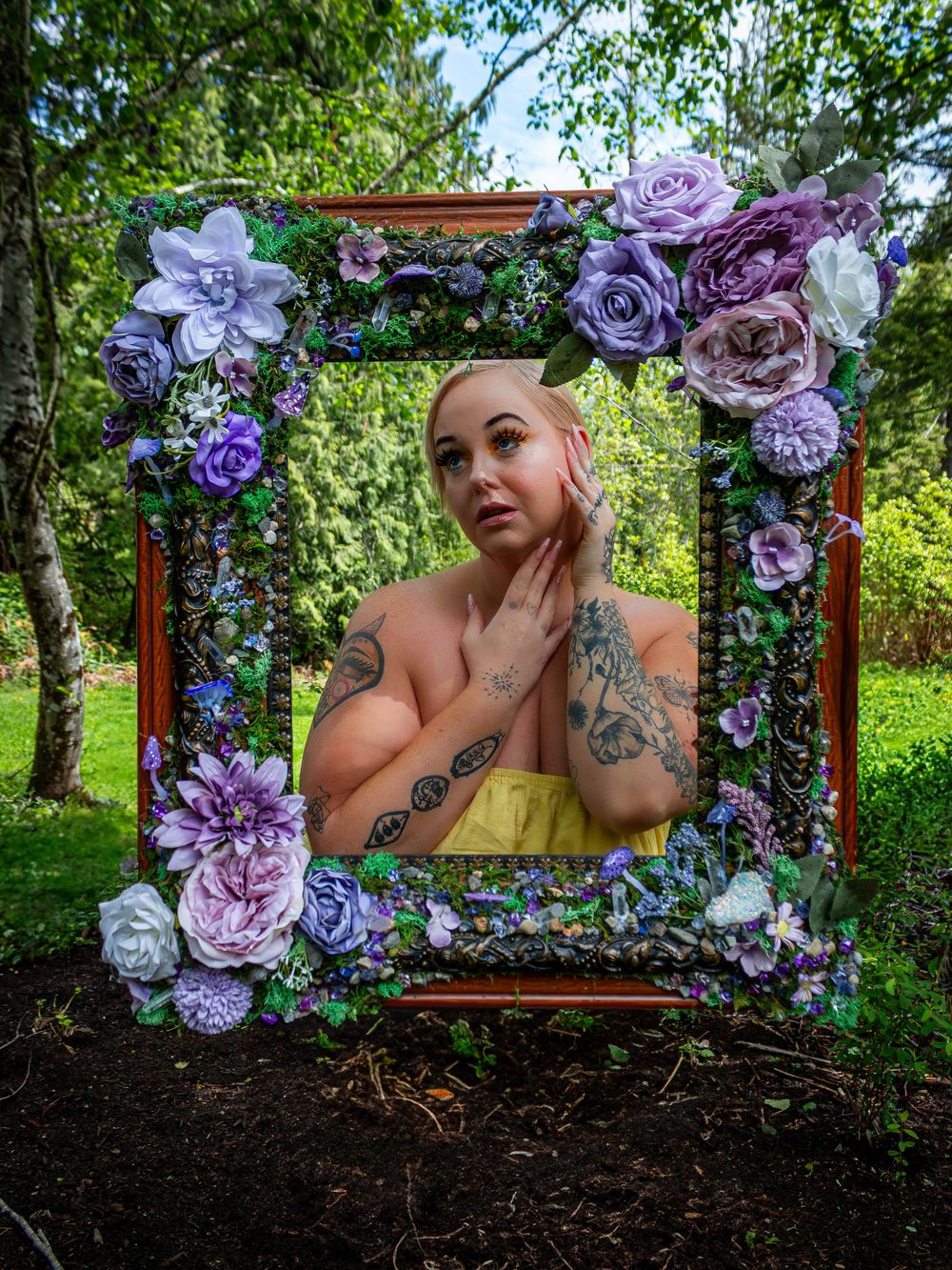 Large wooden frame adorned with purple and white paper flowers creates an artistic outdoor portrait setting in a garden.