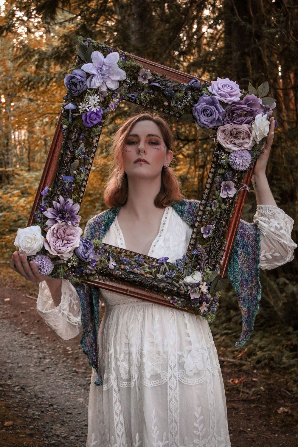 A decorated vintage wooden frame with purple and pink flowers held up in a dreamy forest setting at golden hour.