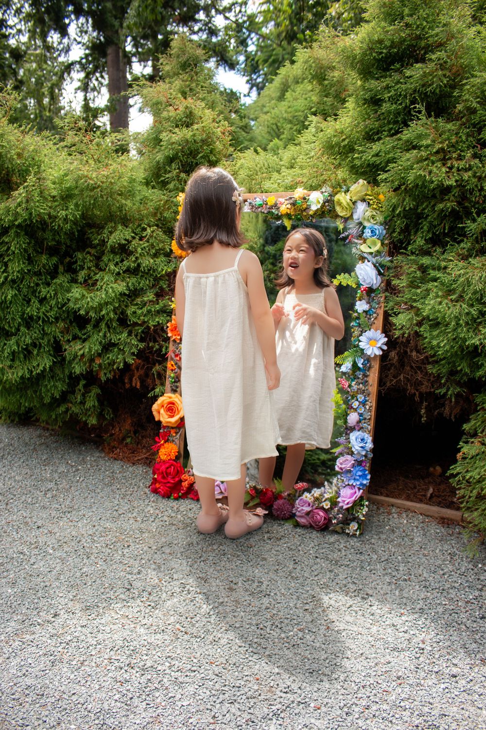 A little girl in a white dress looks at her reflection, which, surprisingly, is doing something different than she is!