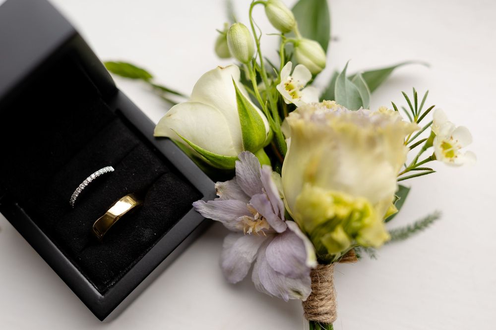 Wedding rings in black box with delicate floral boutonniere featuring white roses and lavender blooms.