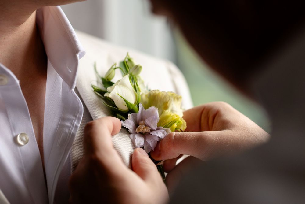 Hands pin a delicate white rose and lavender floral boutonniere onto a light-colored dress shirt.