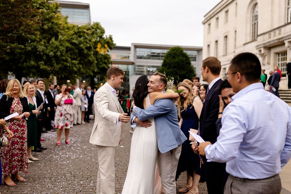 Wedding party and guests gather outside historic building during daytime celebration.